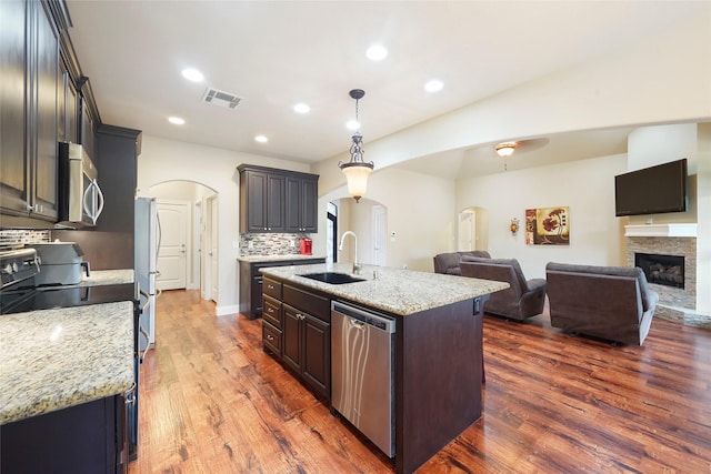 kitchen with dark hardwood / wood-style flooring, stainless steel appliances, sink, a stone fireplace, and an island with sink