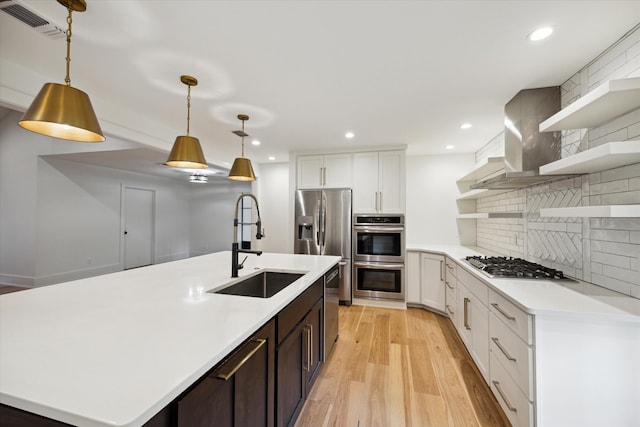 kitchen featuring wall chimney range hood, stainless steel appliances, decorative light fixtures, white cabinets, and light hardwood / wood-style flooring