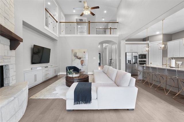 living room with a towering ceiling, a stone fireplace, light wood-type flooring, and ceiling fan