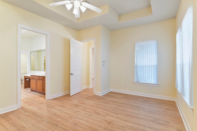 unfurnished bedroom featuring a raised ceiling, ensuite bathroom, ceiling fan, and light wood-type flooring