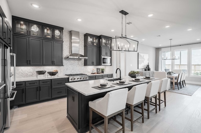 kitchen featuring wall chimney range hood, light hardwood / wood-style flooring, appliances with stainless steel finishes, and a kitchen island with sink