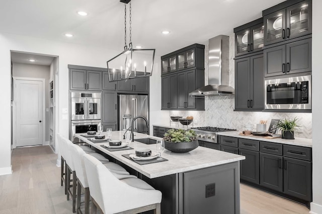 kitchen featuring wall chimney range hood, an island with sink, light hardwood / wood-style flooring, sink, and appliances with stainless steel finishes