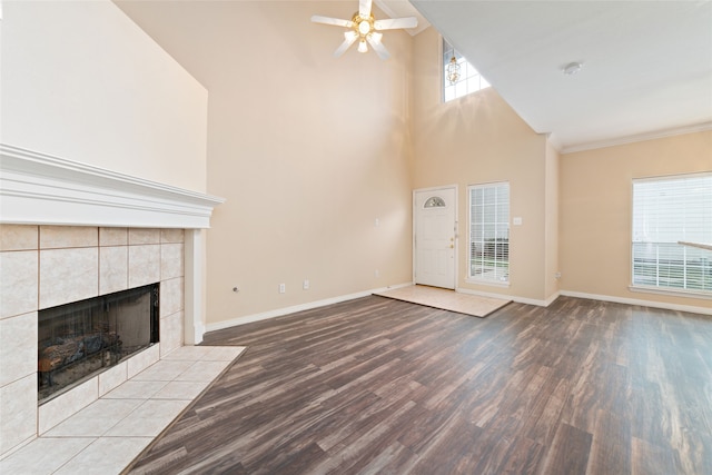 unfurnished living room featuring a tiled fireplace, ceiling fan, a towering ceiling, light hardwood / wood-style floors, and crown molding