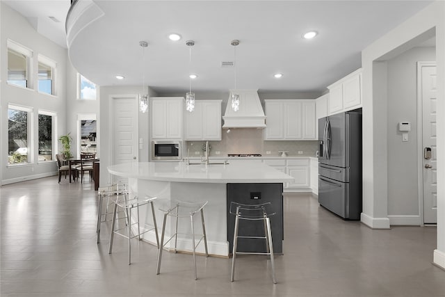 kitchen featuring an island with sink, appliances with stainless steel finishes, white cabinetry, custom range hood, and pendant lighting