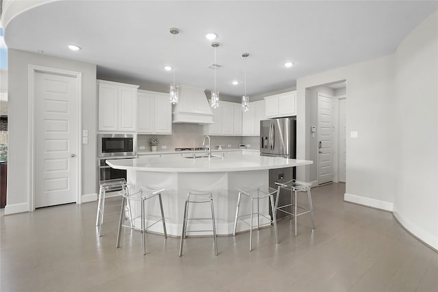 kitchen featuring a kitchen island with sink, custom range hood, hanging light fixtures, white cabinets, and appliances with stainless steel finishes