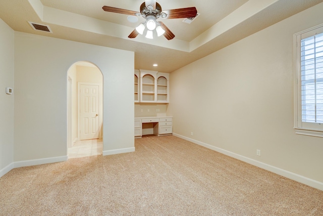 unfurnished room with a ceiling fan, visible vents, built in study area, a raised ceiling, and light carpet