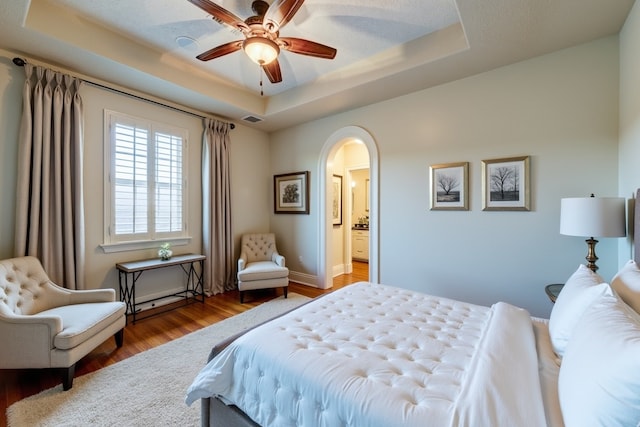 bedroom featuring a tray ceiling, wood-type flooring, ensuite bath, and ceiling fan