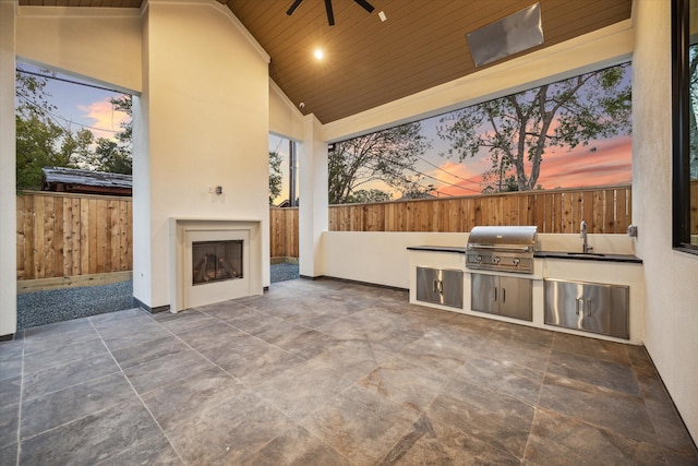 patio terrace at dusk with ceiling fan, exterior kitchen, sink, grilling area, and an outdoor fireplace