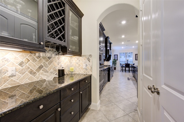 kitchen with dark brown cabinets, dark stone countertops, light tile patterned floors, and backsplash