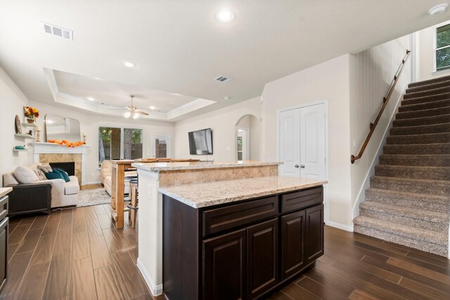 kitchen featuring dark brown cabinets, a tray ceiling, ceiling fan, a kitchen island, and a tiled fireplace