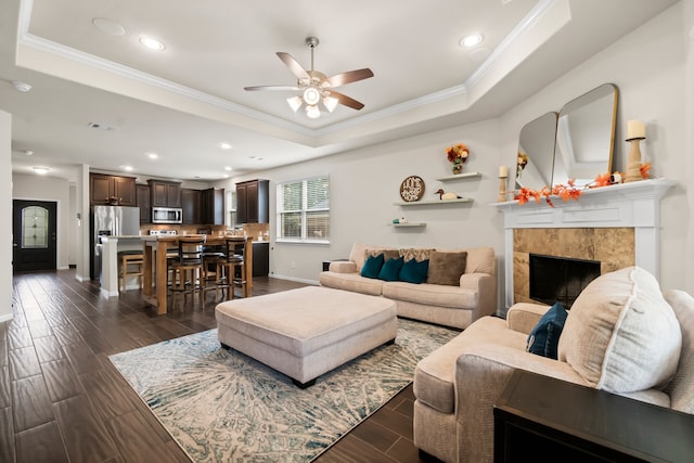 living room featuring dark wood-type flooring, a raised ceiling, ceiling fan, ornamental molding, and a tiled fireplace