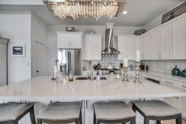 kitchen featuring wall chimney range hood, an island with sink, appliances with stainless steel finishes, white cabinetry, and sink