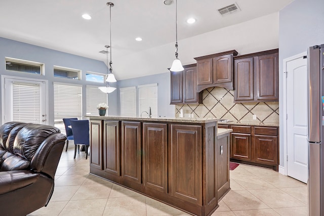 kitchen with a kitchen island with sink, dark brown cabinets, light stone countertops, light tile patterned flooring, and decorative light fixtures