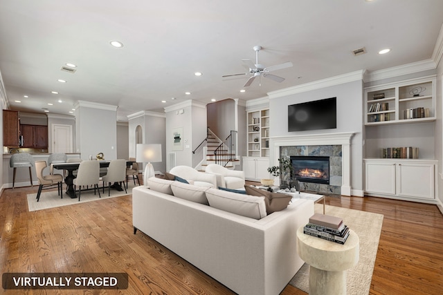 living room featuring ceiling fan, crown molding, light hardwood / wood-style flooring, built in features, and a tiled fireplace