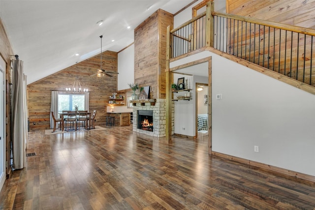 unfurnished living room featuring a stone fireplace, high vaulted ceiling, dark hardwood / wood-style floors, wood walls, and ceiling fan with notable chandelier