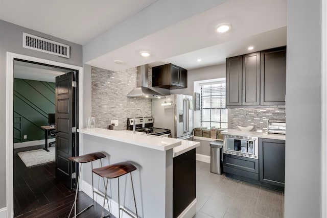 kitchen with wall chimney exhaust hood, kitchen peninsula, stainless steel appliances, a kitchen bar, and tasteful backsplash