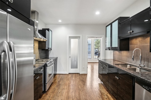 kitchen with wall chimney exhaust hood, stainless steel appliances, dark stone countertops, sink, and light wood-type flooring