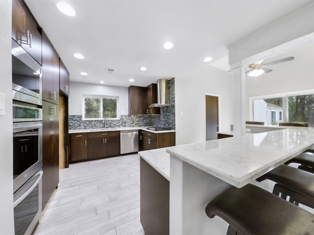 kitchen featuring wall chimney range hood, appliances with stainless steel finishes, light wood-type flooring, kitchen peninsula, and a breakfast bar area