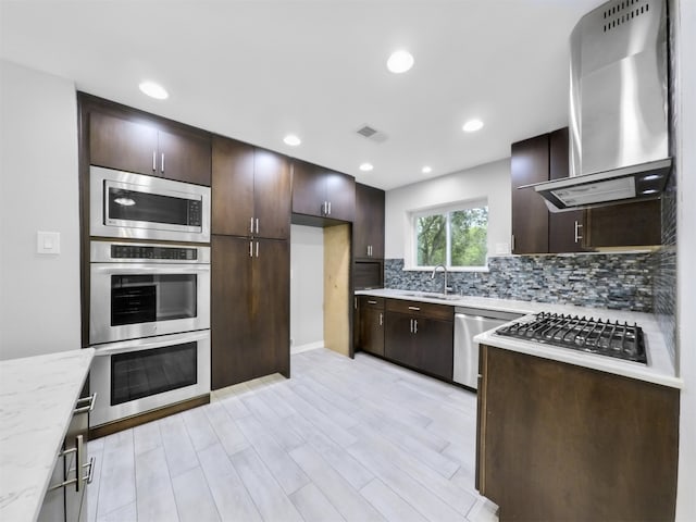 kitchen with wall chimney range hood, light stone counters, light wood-type flooring, sink, and stainless steel appliances