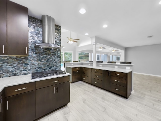 kitchen featuring tasteful backsplash, wall chimney range hood, dark brown cabinets, stainless steel gas stovetop, and light hardwood / wood-style flooring