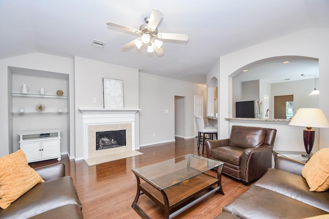 living room featuring a tile fireplace, ceiling fan, built in features, wood-type flooring, and lofted ceiling