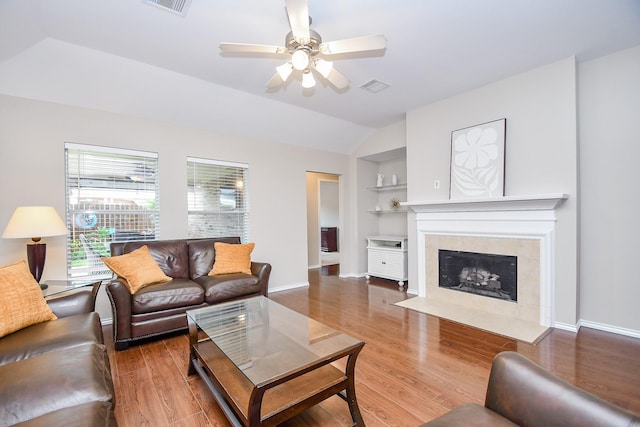 living room with built in shelves, ceiling fan, wood-type flooring, lofted ceiling, and a tiled fireplace