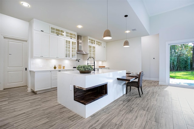 kitchen with decorative backsplash, wall chimney exhaust hood, decorative light fixtures, white cabinetry, and an island with sink