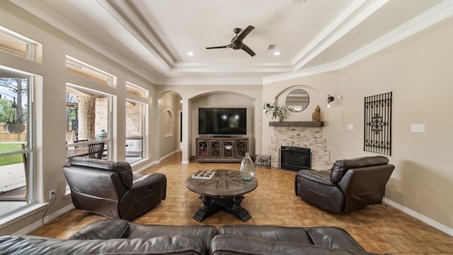 living room featuring a tray ceiling, a stone fireplace, ceiling fan, and crown molding