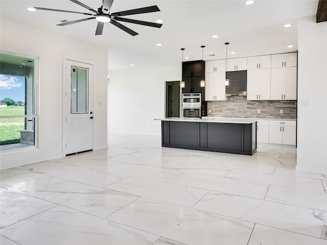 kitchen with decorative backsplash, white cabinets, an island with sink, pendant lighting, and double oven