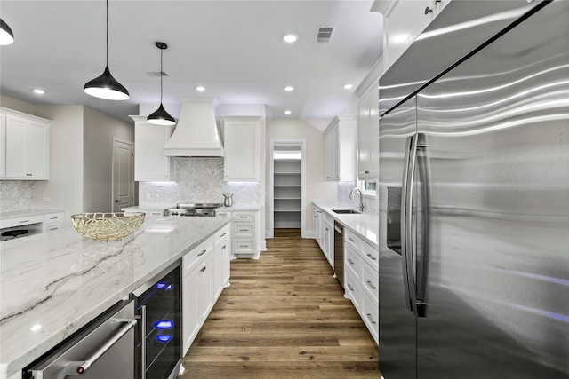 kitchen with premium range hood, stainless steel refrigerator with ice dispenser, hanging light fixtures, white cabinets, and beverage cooler