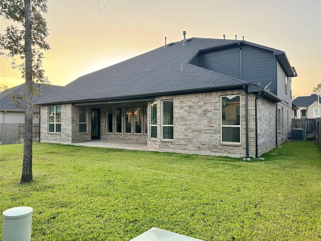 back of house at dusk featuring brick siding, a yard, a patio, a shingled roof, and fence