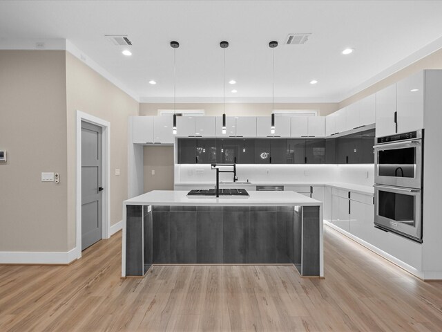 kitchen with decorative light fixtures, light wood-type flooring, white cabinetry, an island with sink, and stainless steel double oven