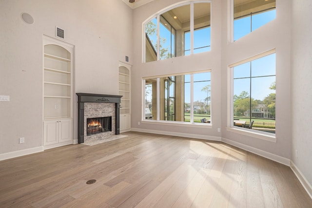 unfurnished living room with a towering ceiling, a stone fireplace, wood-type flooring, and built in shelves