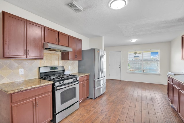 kitchen with appliances with stainless steel finishes, backsplash, a textured ceiling, light stone counters, and light hardwood / wood-style flooring