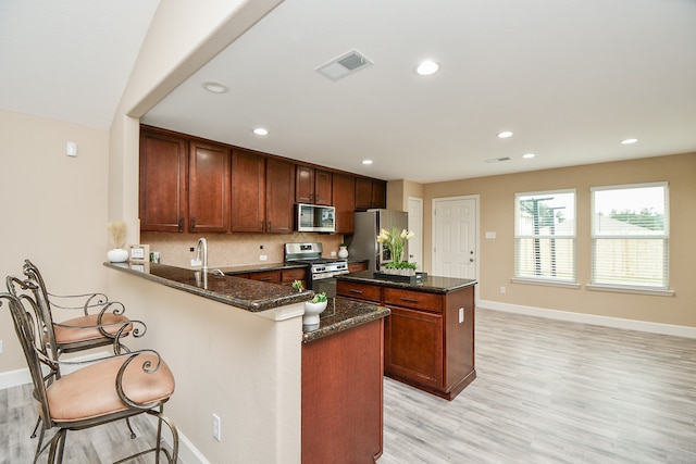 kitchen with appliances with stainless steel finishes, kitchen peninsula, a breakfast bar area, dark stone counters, and light hardwood / wood-style flooring