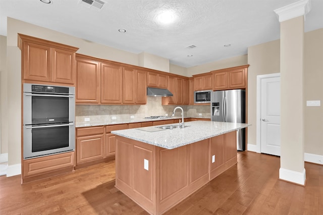 kitchen featuring tasteful backsplash, light stone counters, a center island with sink, stainless steel appliances, and light hardwood / wood-style flooring