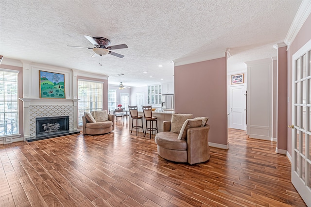 living room with a tiled fireplace, a textured ceiling, hardwood / wood-style flooring, ornamental molding, and ceiling fan