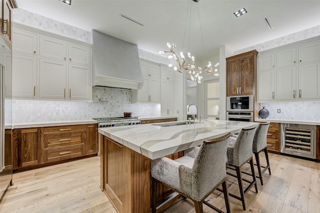 kitchen with wine cooler, premium range hood, a kitchen island with sink, light wood-type flooring, and sink