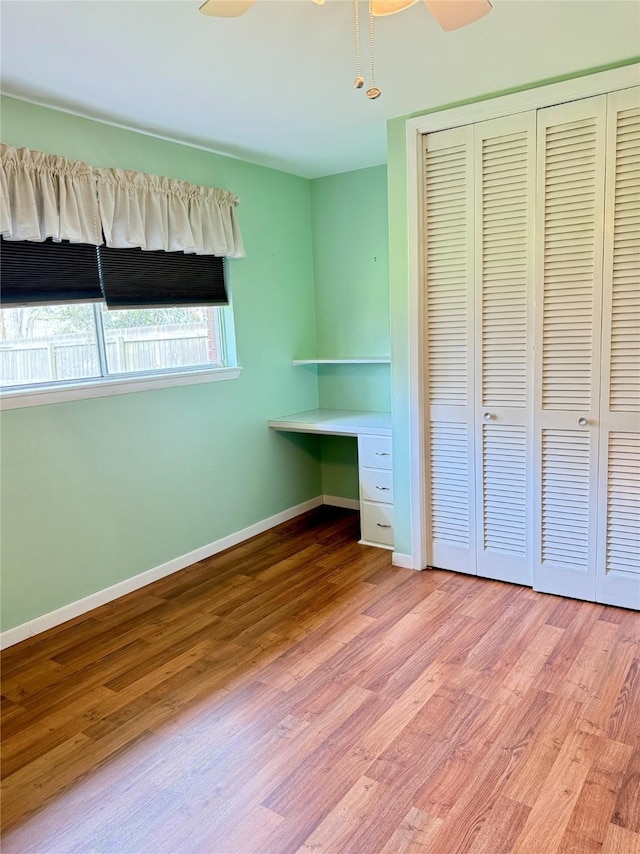 unfurnished bedroom with a closet, a ceiling fan, wood finished floors, built in study area, and baseboards