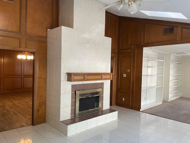 unfurnished living room featuring a tiled fireplace, light colored carpet, wood walls, ceiling fan, and built in features