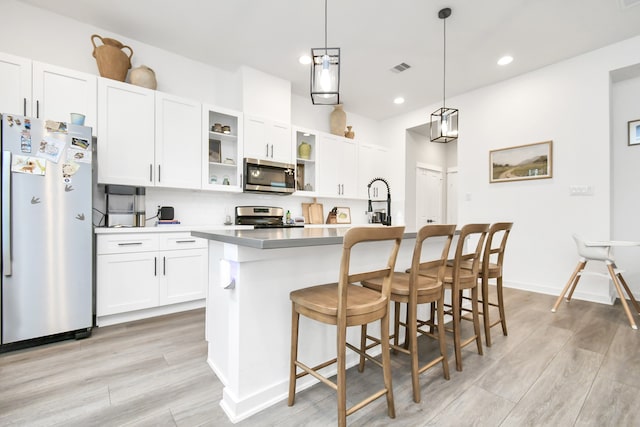 kitchen with stainless steel appliances, white cabinetry, decorative light fixtures, an island with sink, and light hardwood / wood-style floors