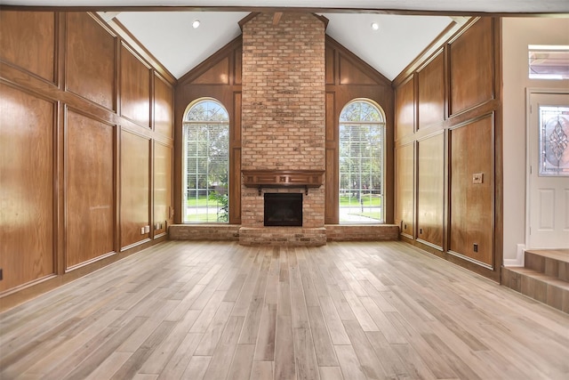 unfurnished living room featuring wooden walls, a brick fireplace, high vaulted ceiling, and light hardwood / wood-style floors