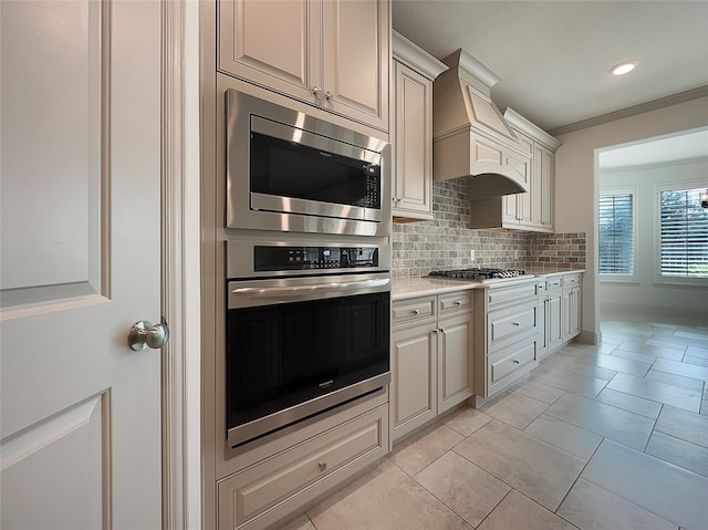 kitchen with appliances with stainless steel finishes, ornamental molding, tasteful backsplash, light tile patterned floors, and custom exhaust hood