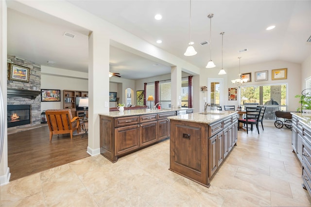 kitchen featuring a kitchen island with sink, a fireplace, decorative light fixtures, light stone countertops, and ceiling fan with notable chandelier