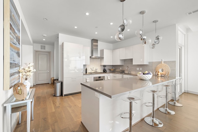 kitchen with a kitchen breakfast bar, decorative light fixtures, white cabinets, wall chimney range hood, and light wood-type flooring