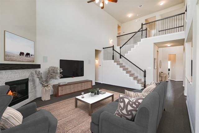 living room featuring ceiling fan, a towering ceiling, a stone fireplace, and dark hardwood / wood-style flooring