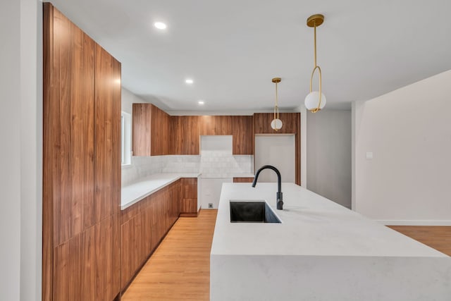 kitchen with sink, tasteful backsplash, decorative light fixtures, a center island with sink, and light wood-type flooring