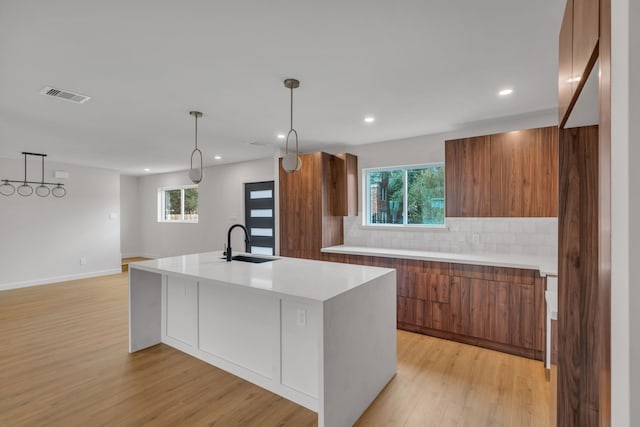 kitchen with light wood-type flooring, backsplash, sink, pendant lighting, and an island with sink