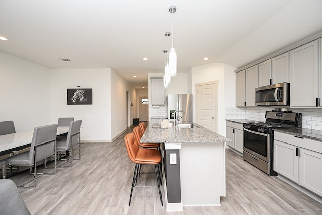 kitchen with light hardwood / wood-style floors, an island with sink, light stone countertops, pendant lighting, and appliances with stainless steel finishes