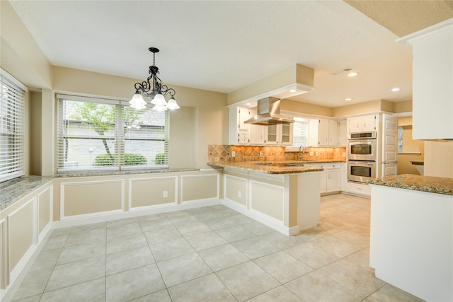 kitchen featuring kitchen peninsula, decorative light fixtures, double oven, white cabinets, and island exhaust hood